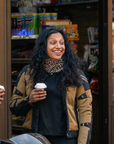 A woman smiling and holding a takeaway coffee cup stands in front of a convenience store entrance. She is wearing a tan and black motorcycle-style jacket over a black top and a patterned scarf.