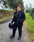 a smiling red haired woman motorcyclist wearing witches of wheels t-shirt, black jeans and mc jacket