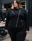 Woman wearing a black jacket with zippers standing in front of a motorcycle.
