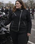 Woman in black motorcycle gear standing next to a motorcycle in a parking lot.