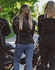 Three women in black motorcycle jackets standing next to motorcycles on a sunny day.