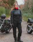 A woman wearing a black motorcycle jacket, black pants, and a bright red neck gaiter stands on a gravel path between two motorcycles in an outdoor setting with autumn trees.