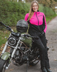 A woman wearing a bright pink and black motorcycle riding jacket, black pants, and gloves stands next to an olive green cruiser motorcycle on a paved path outdoors.