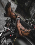 women's feet resting on motorcycle steering wheel, wearing brown leather mc boots
