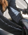 Close-up of a rider wearing black and gray Shimaa arm warmers with white text reading 'HITTING THE LIMITS // SHIMAA' on the forearm, along with black gloves and a motorcycle handlebar in the background.