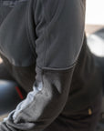 Close-up of a person wearing a dark gray long-sleeve quarter-zip fleece jacket, leaning over a black motorcycle in an outdoor setting.