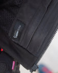 Close-up of a black technical jacket or vest showing the inner mesh lining, a zipper, and a small sewn label with white text.