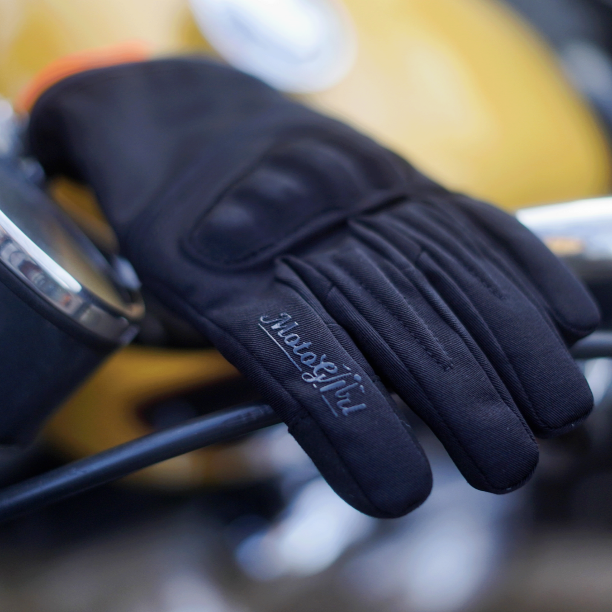 BLACK MOTORCYCLE GLOVES WITH MOTOGIRL LOGO ON A HANDLE BAR OF A MOTORCYCLE