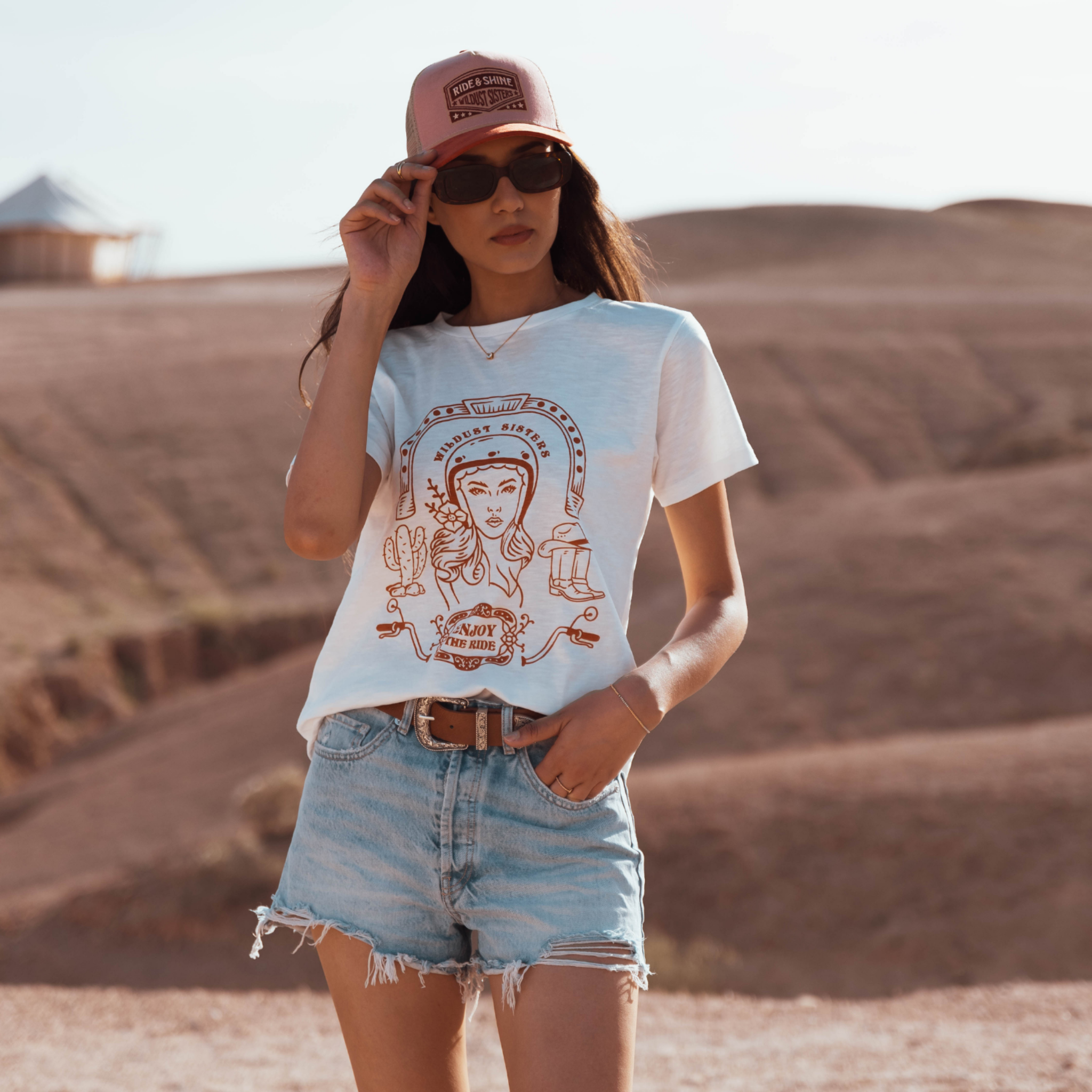 Woman in the dessert wearing a white T-shirt with a Woman wearing a Motorcyclehelmet print and the text "Wild dust sister" and "Enjoy The ride" in orange