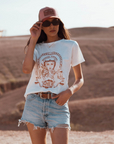 Woman in the dessert wearing a white T-shirt with a Woman wearing a Motorcyclehelmet print and the text "Wild dust sister" and "Enjoy The ride" in orange