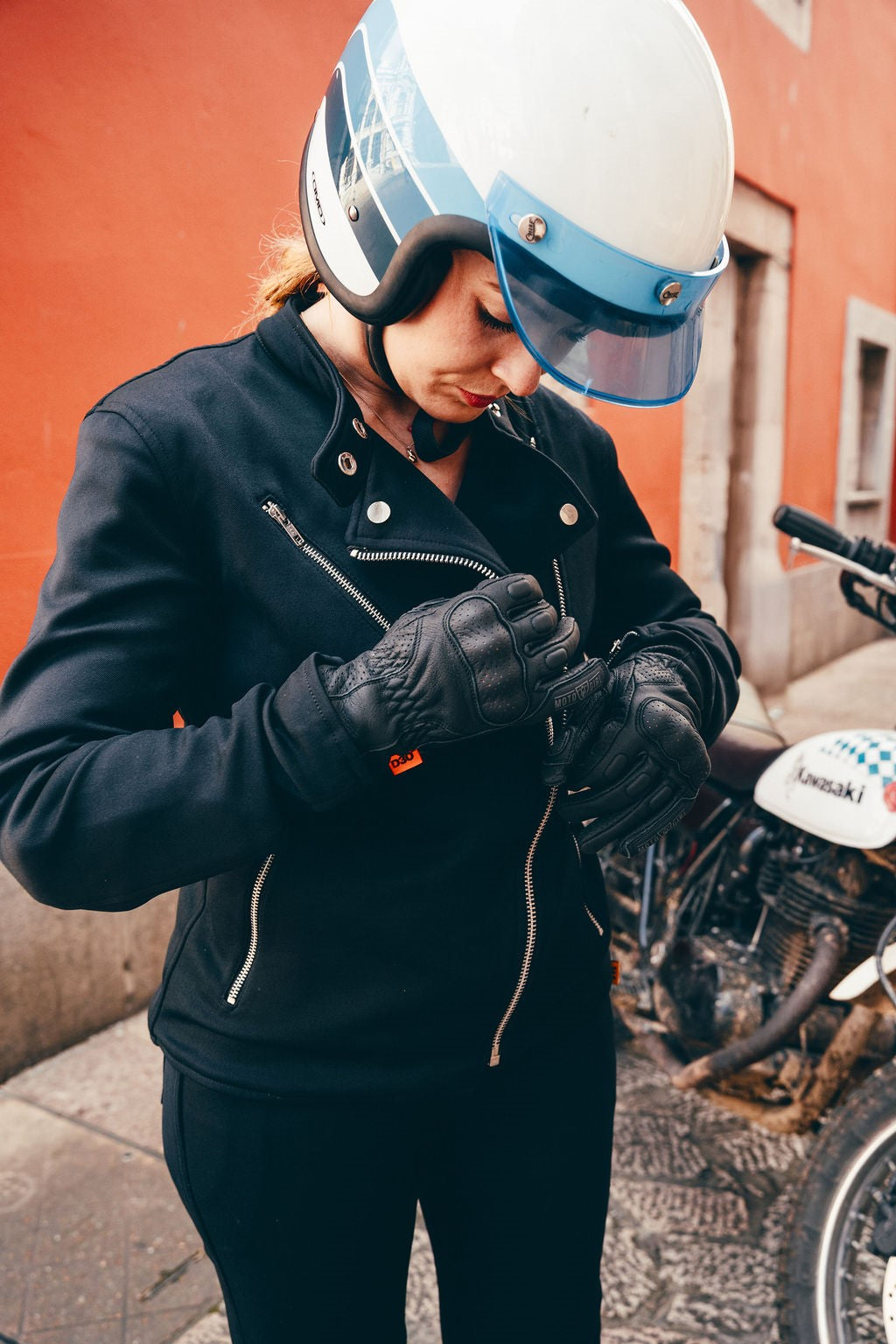 Person wearing a black jacket and helmet, standing next to a motorcycle with a red wall in the background.