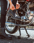 a woman standing by her motorcycle and zipping her black leather boot 