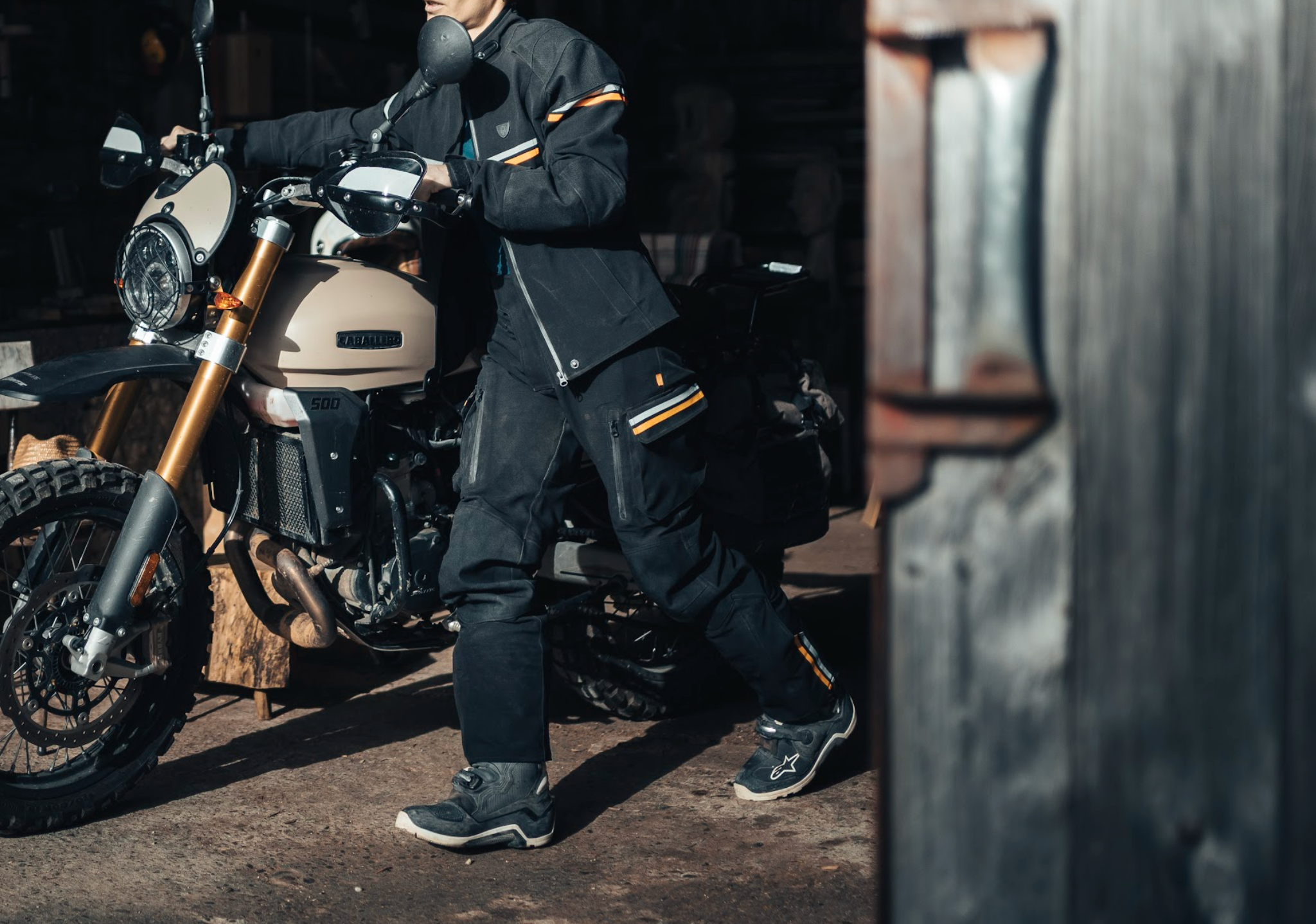 Person in dark clothing with gold accents standing next to a motorcycle in a rustic setting