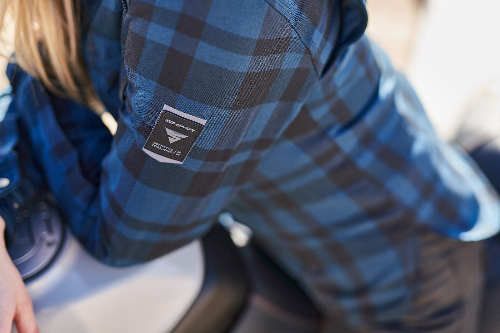 Person wearing a blue plaid shirt with a SHIMA motorcycle wear brand logo on the sleeve