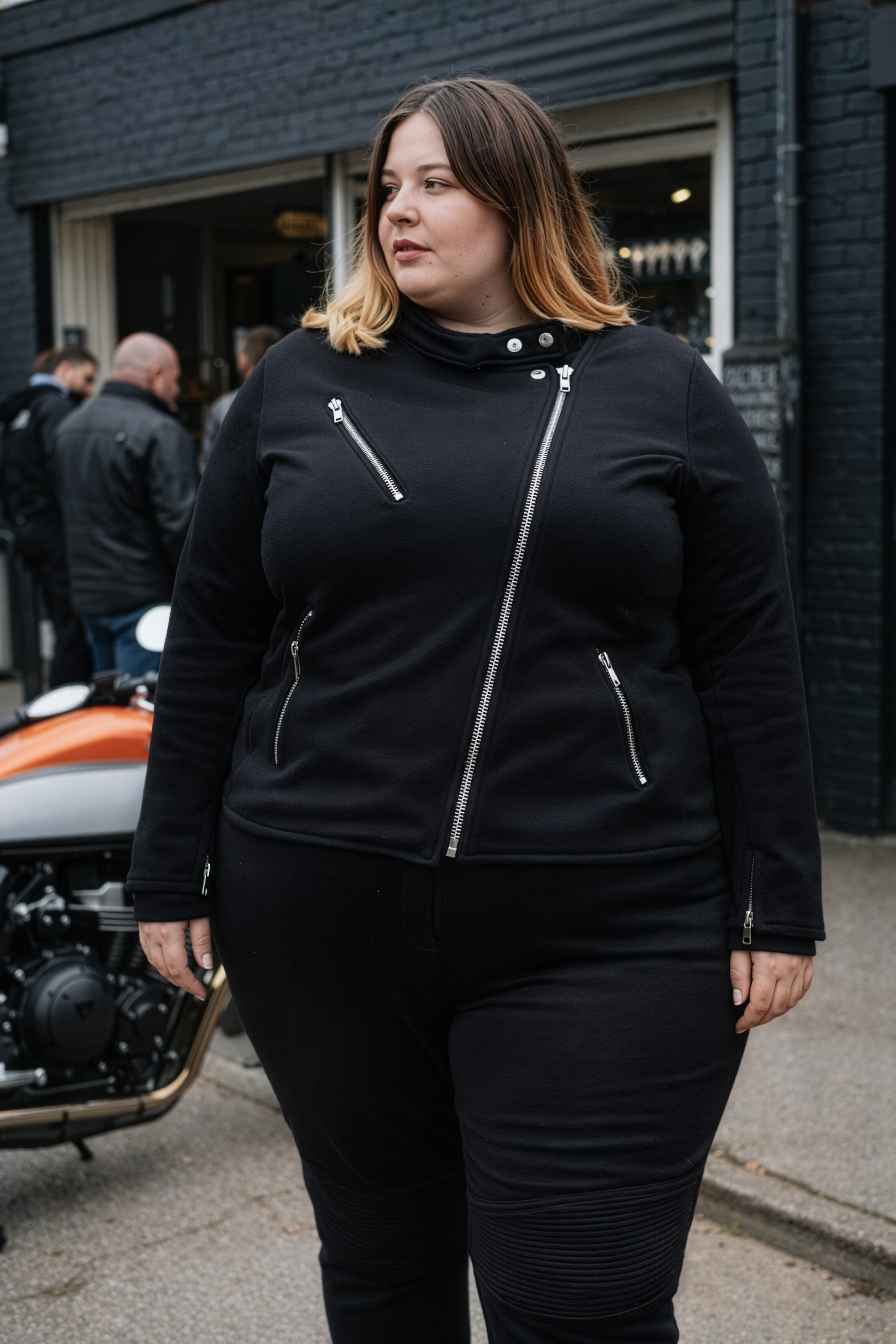 Woman wearing a black jacket with zippers standing in front of a motorcycle.