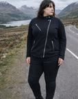 Person wearing a black outfit standing on a road with mountains and a lake in the background
