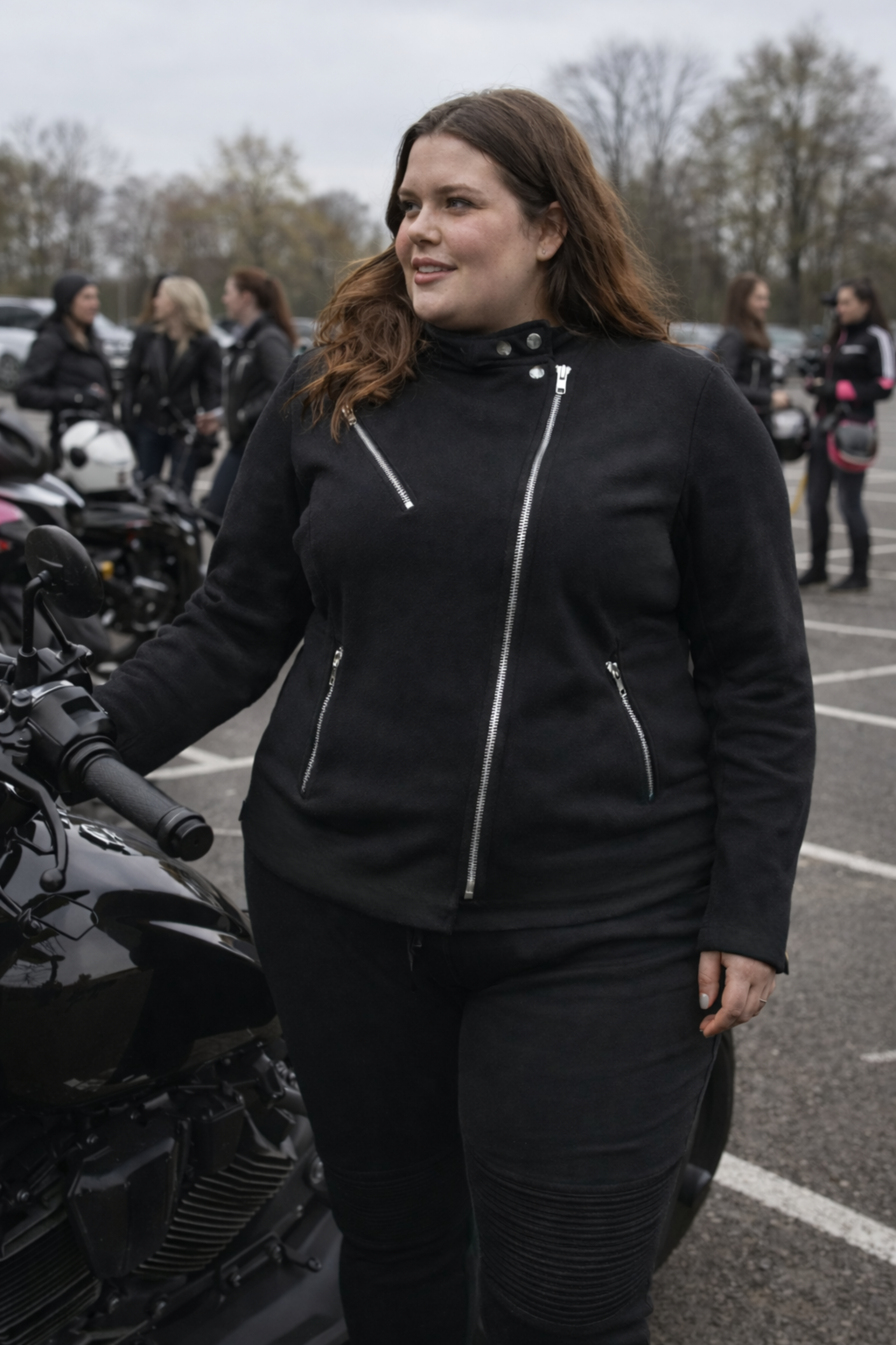 Woman in black motorcycle gear standing next to a motorcycle in a parking lot.