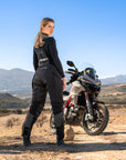 A woman standing by her motorcycle wearing Black SHIMA touring motorcycle pants for women