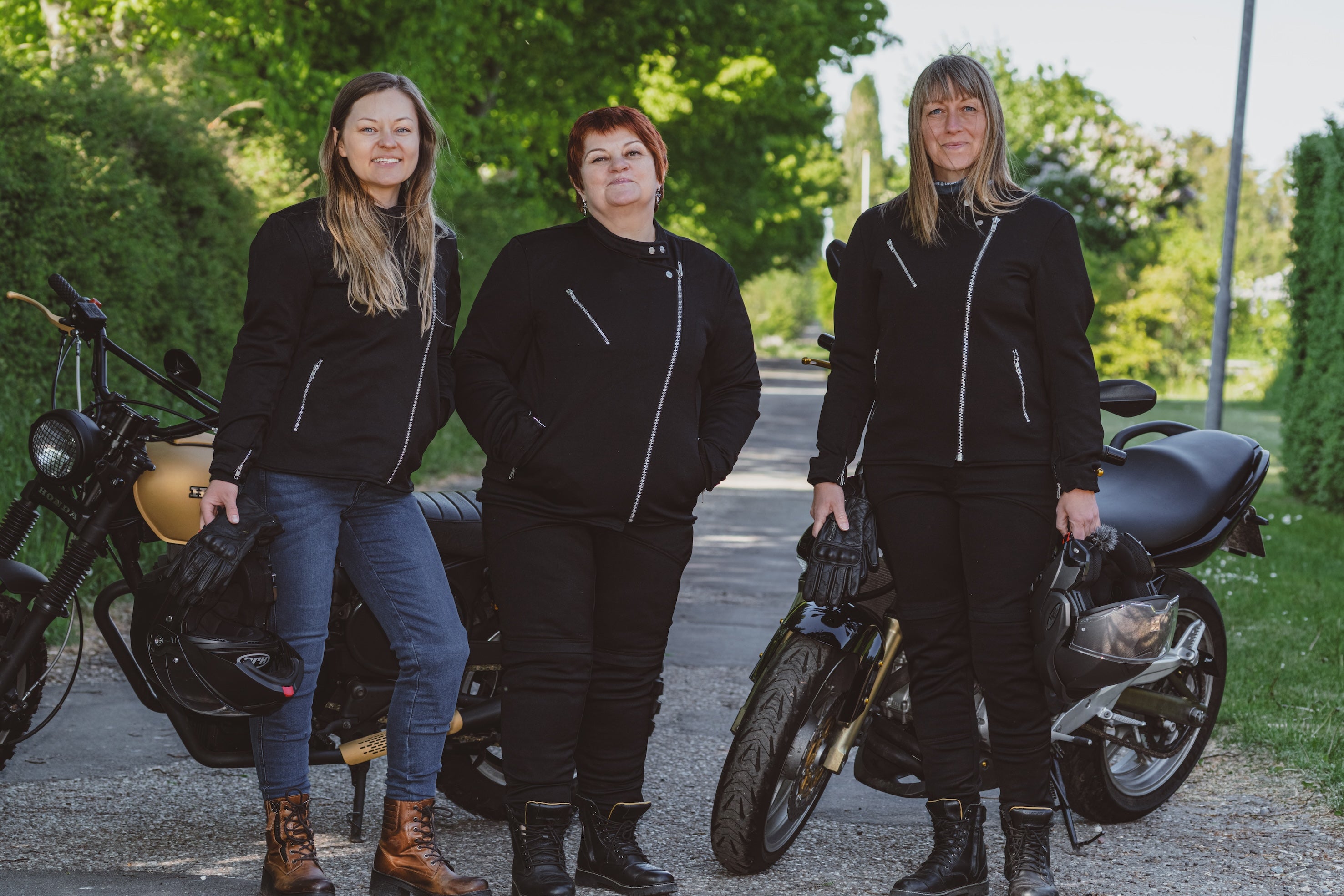 Three women standing next to motorcycles on a road with trees in the background