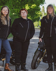 Three women standing next to motorcycles on a road with trees in the background