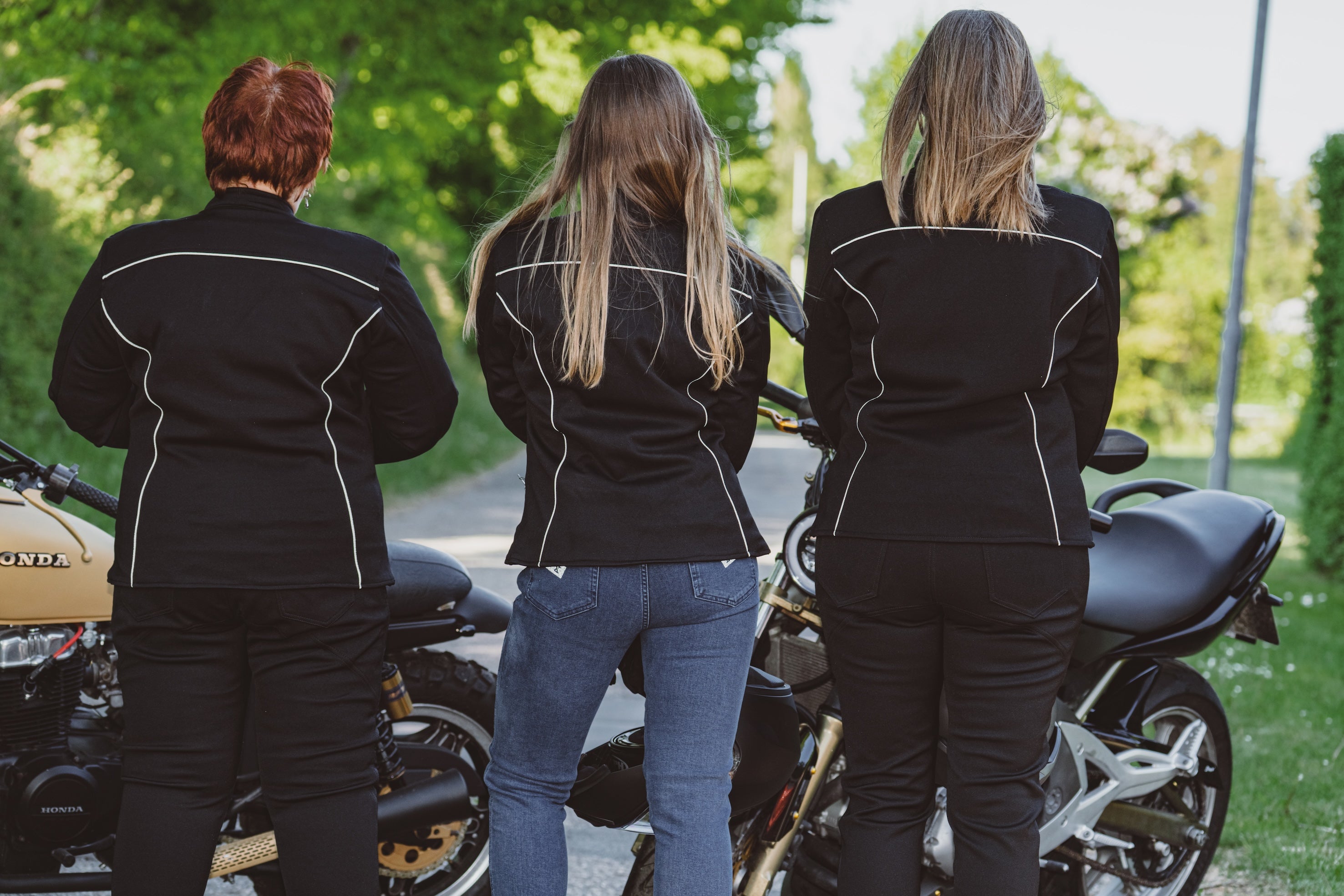 Three women in black motorcycle jackets standing next to motorcycles on a sunny day.