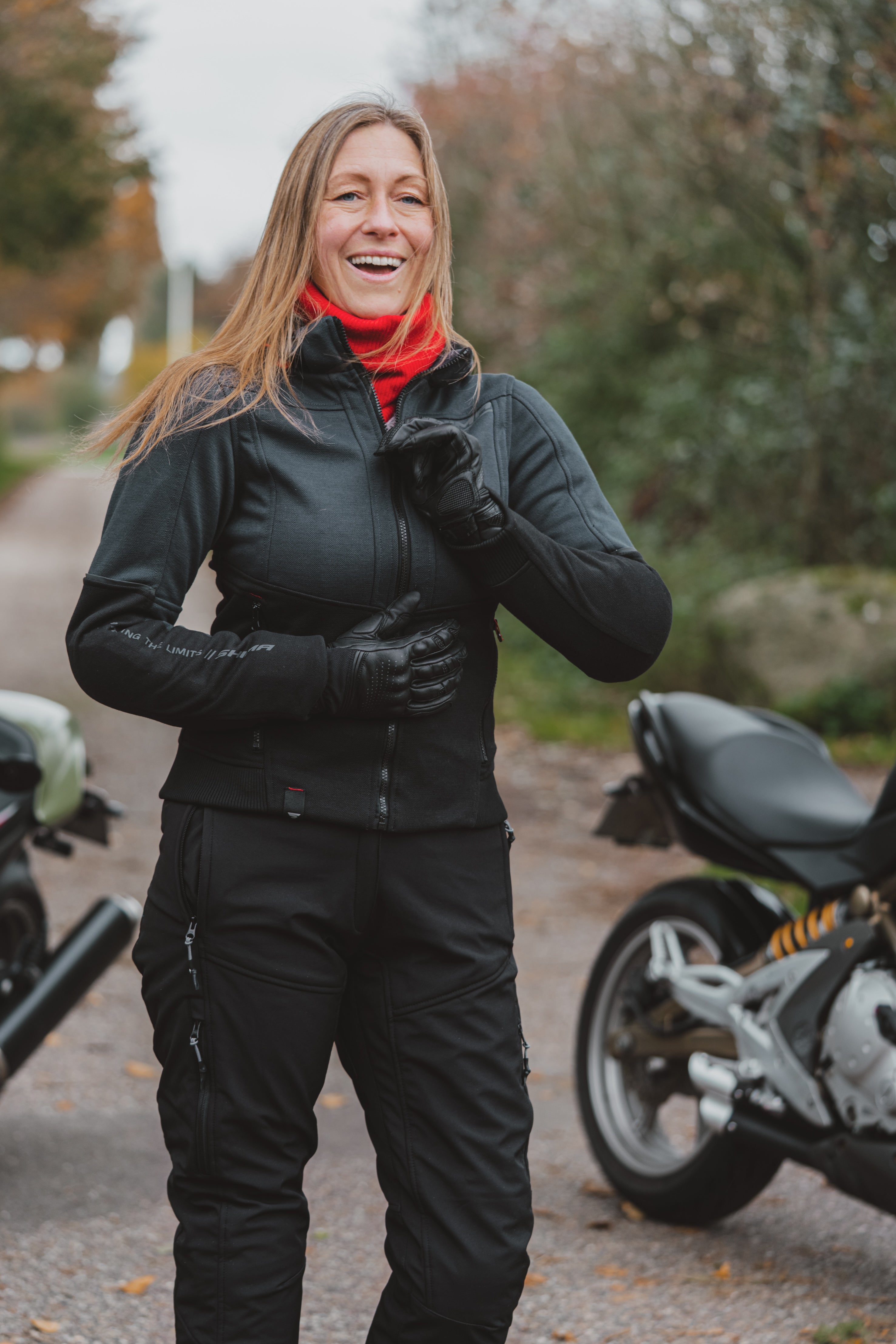 A woman wearing a black motorcycle jacket and matching pants with a bright red neck gaiter stands outdoors on a path with motorcycles partially visible on both sides.