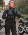 A woman wearing a black motorcycle jacket and matching pants with a bright red neck gaiter stands outdoors on a path with motorcycles partially visible on both sides.