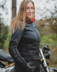 A woman wearing a black motorcycle jacket, matching black pants, and black gloves, standing outdoors next to a motorcycle with a blurred forest background.