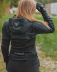 A woman viewed from behind wearing a black SHIMA insulated jacket and matching black pants, standing outdoors on a grassy path.