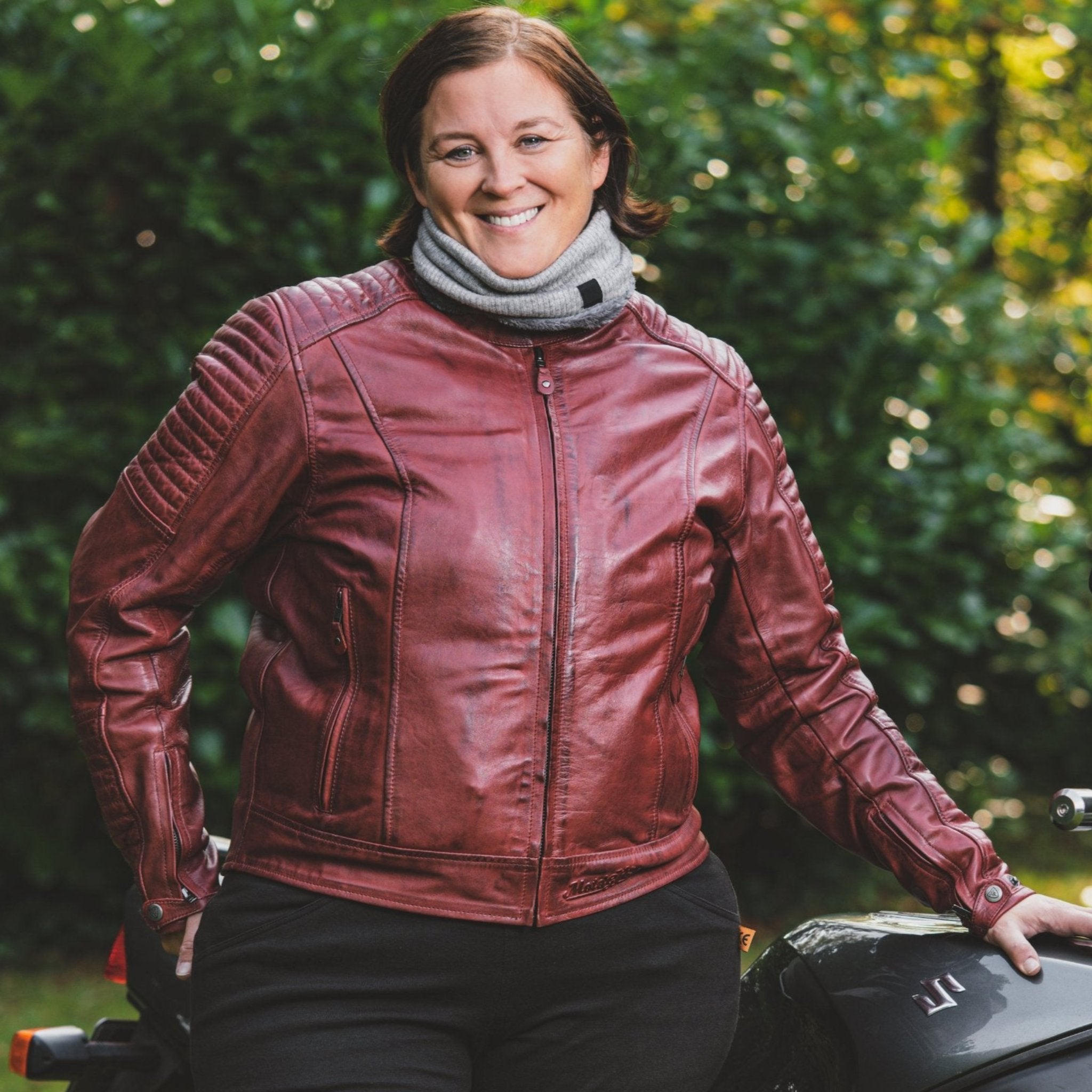 Woman in a red Valerie jacket and light gray neck warmer standing outside next to a Suzuki motorcycle.