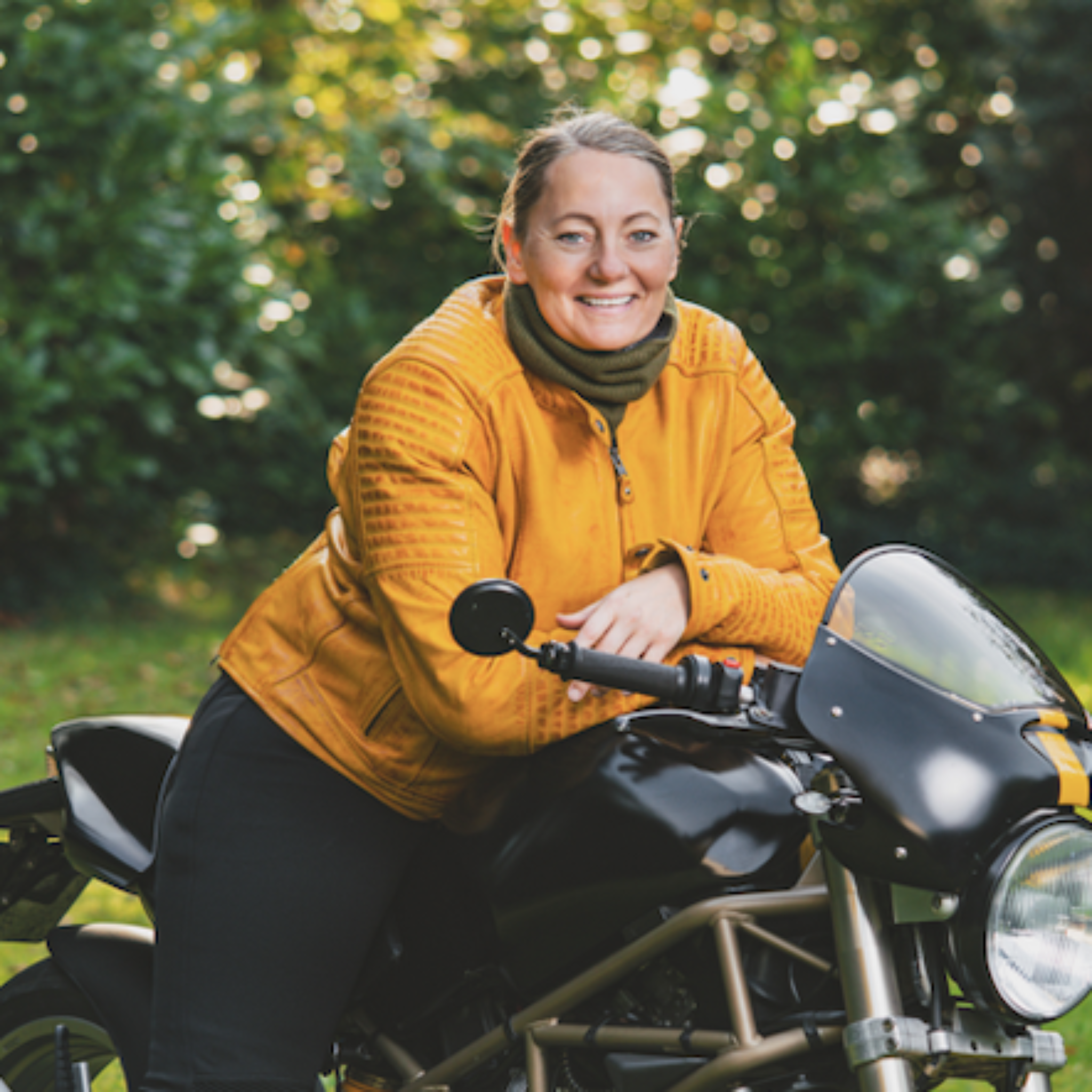 Smiling women sitting on a motorcycle surrounded by nature, wearing a yellow Valerie jacket and a green knitted neck warmer from Moto Girl.