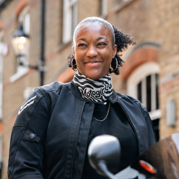 A woman wearing a black motorcycle jacket and a black-and-white patterned neck gaiter with the word 'Motogadget' printed on it, standing outdoors in front of a brick building.