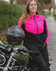 A woman wearing a bright pink and black motorcycle jacket sits on an olive green cruiser motorcycle outdoors. A black helmet rests on the motorcycle seat, and trees and a road are blurred in the background.