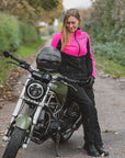 A woman wearing a bright pink and black motorcycle jacket, black pants, and gloves sits on a cruiser-style motorcycle on an outdoor path.