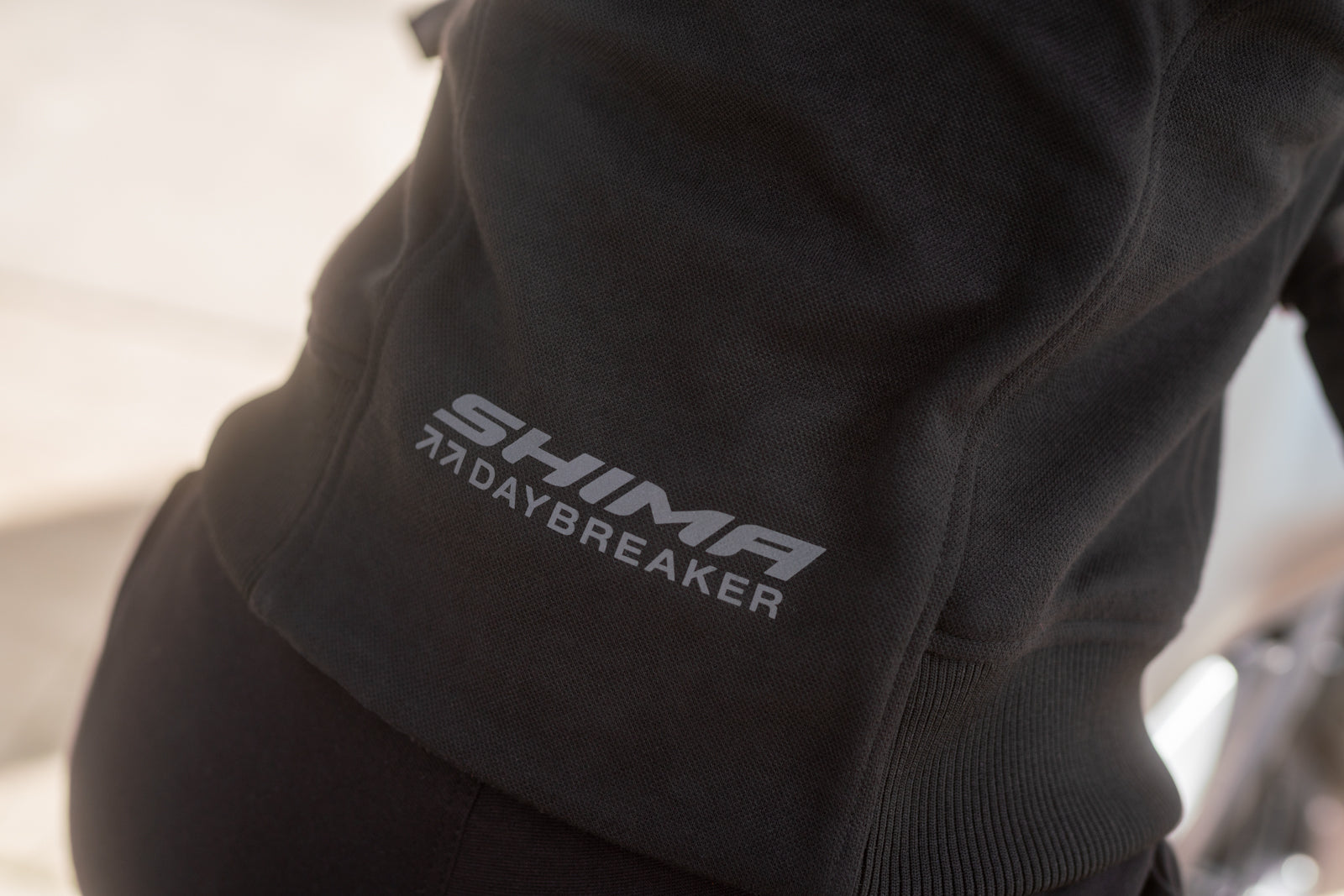Close-up of a black hoodie with a gray embroidered logo reading 'SHIMA 7DAYBREAKER' on the back, worn by a person.